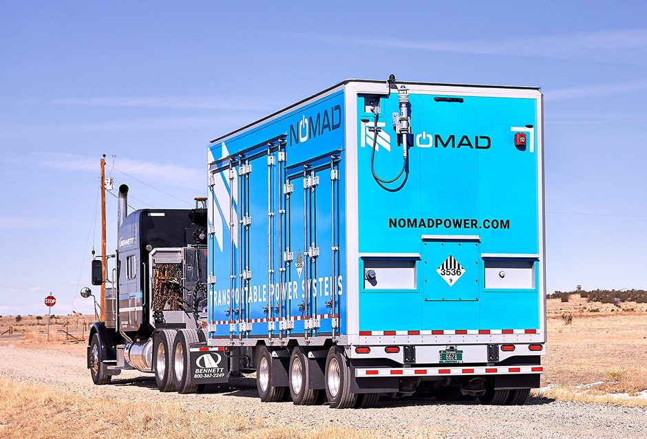 A blue NOMAD Voyager transportable battery unit on a semi-trailer, parked on a gravel road in open country.